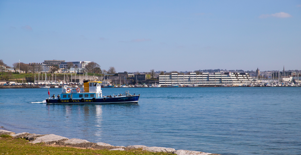 A small blue and yellow passenger ferry moves across a calm harbour on a sunny day, with people seated on deck, rocky shoreline in the foreground, and a backdrop of modern waterfront buildings, green parkland, and moored yachts.
